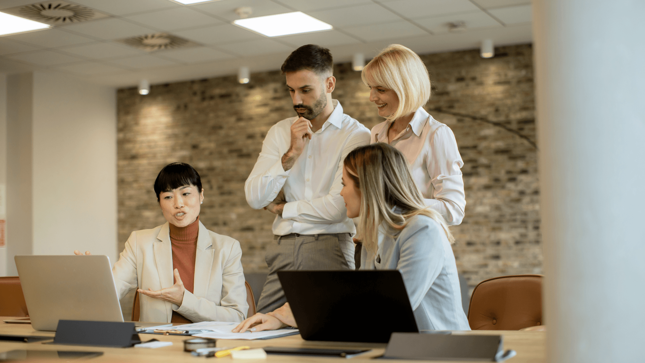 Four colleagues discussing work around a table with laptops, in a modern office setting with a brick wall backdrop.