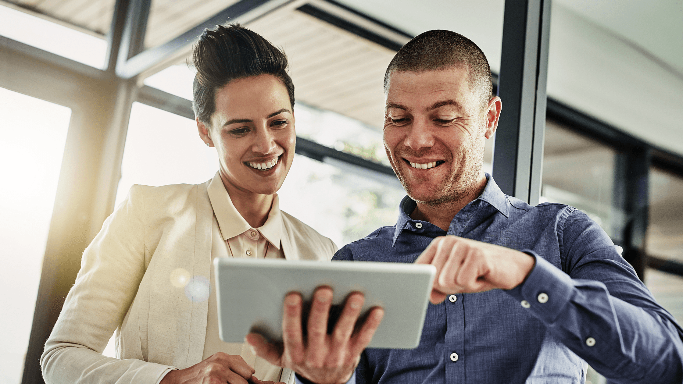 Two people smiling and looking at a tablet in a bright office setting, with large windows in the background.
