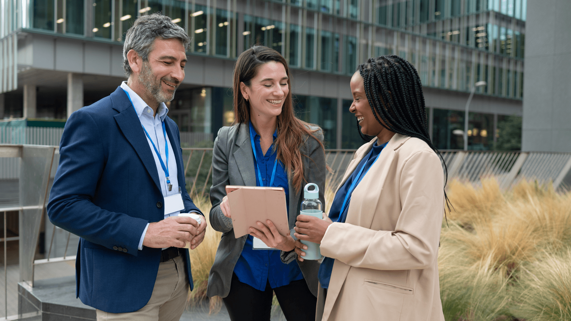 Three professionals standing outside a modern building, smiling and conversing while holding a tablet and a water bottle.