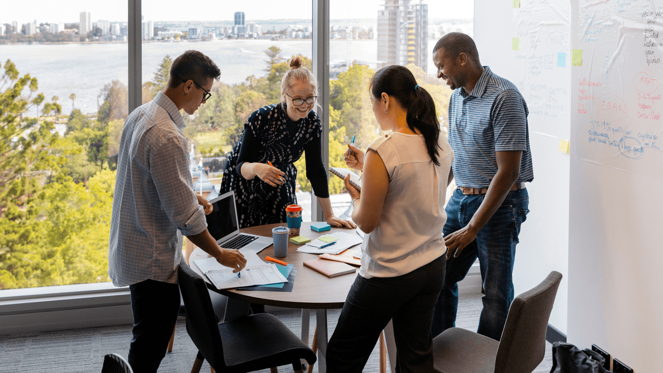 Four people collaborate around a table in an office with large windows showing an urban waterfront view. Notes and diagrams are on a whiteboard.