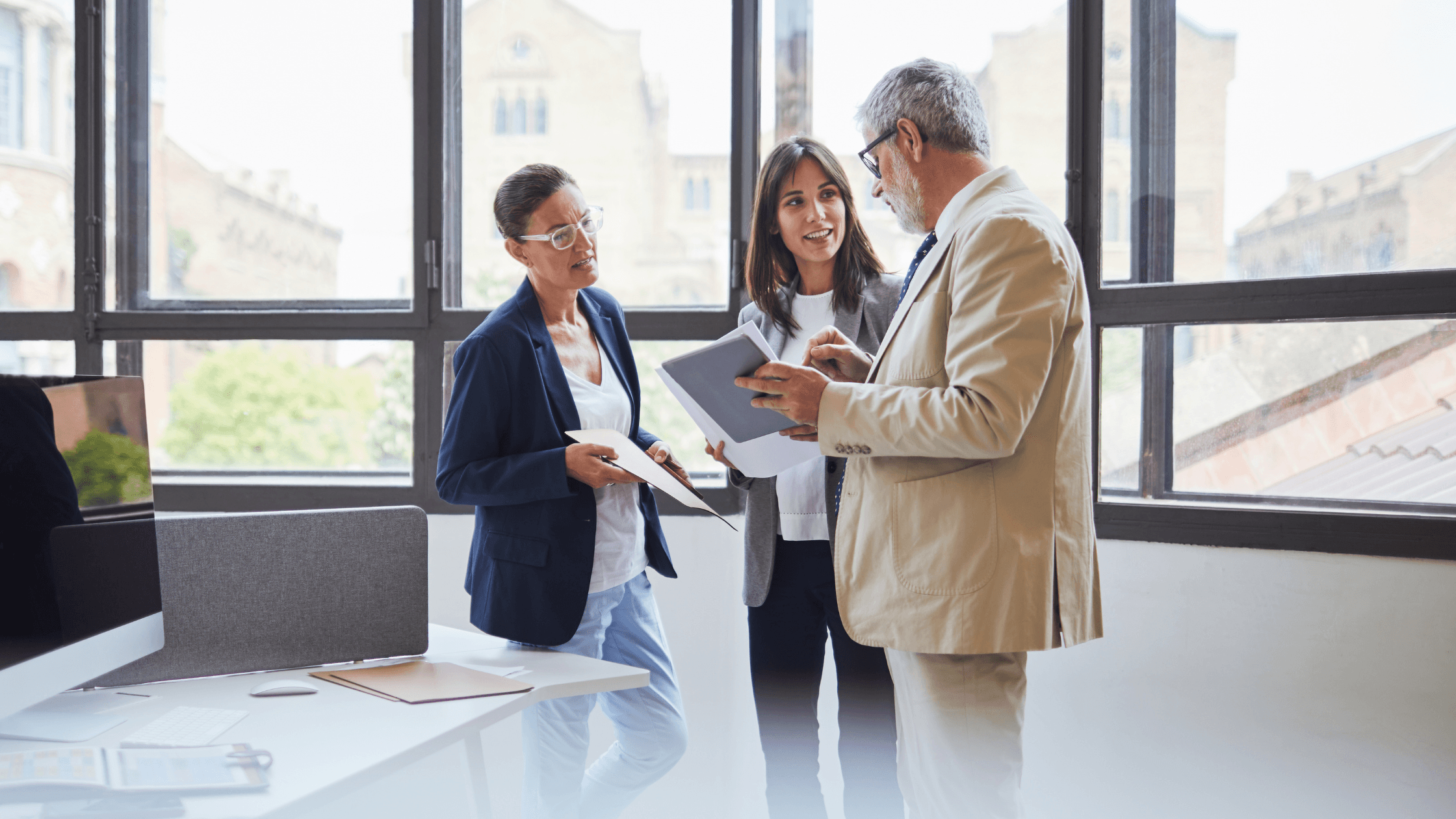 Three professionals in a bright office, discussing papers. Tall windows reveal a blurred view of a building outside.
