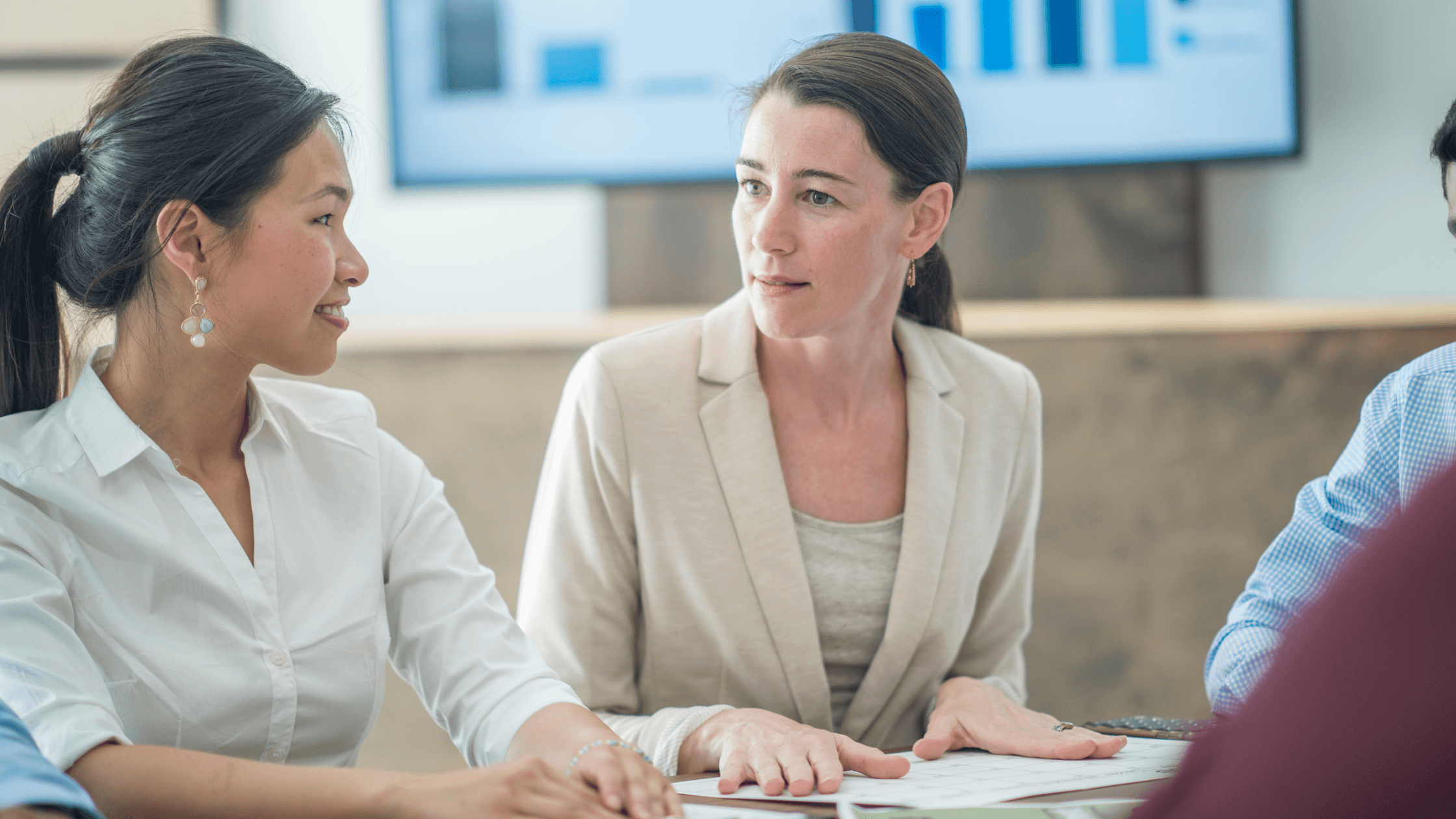 Two women in a business meeting, sitting at a table with documents. A presentation screen with graphs is visible in the background.