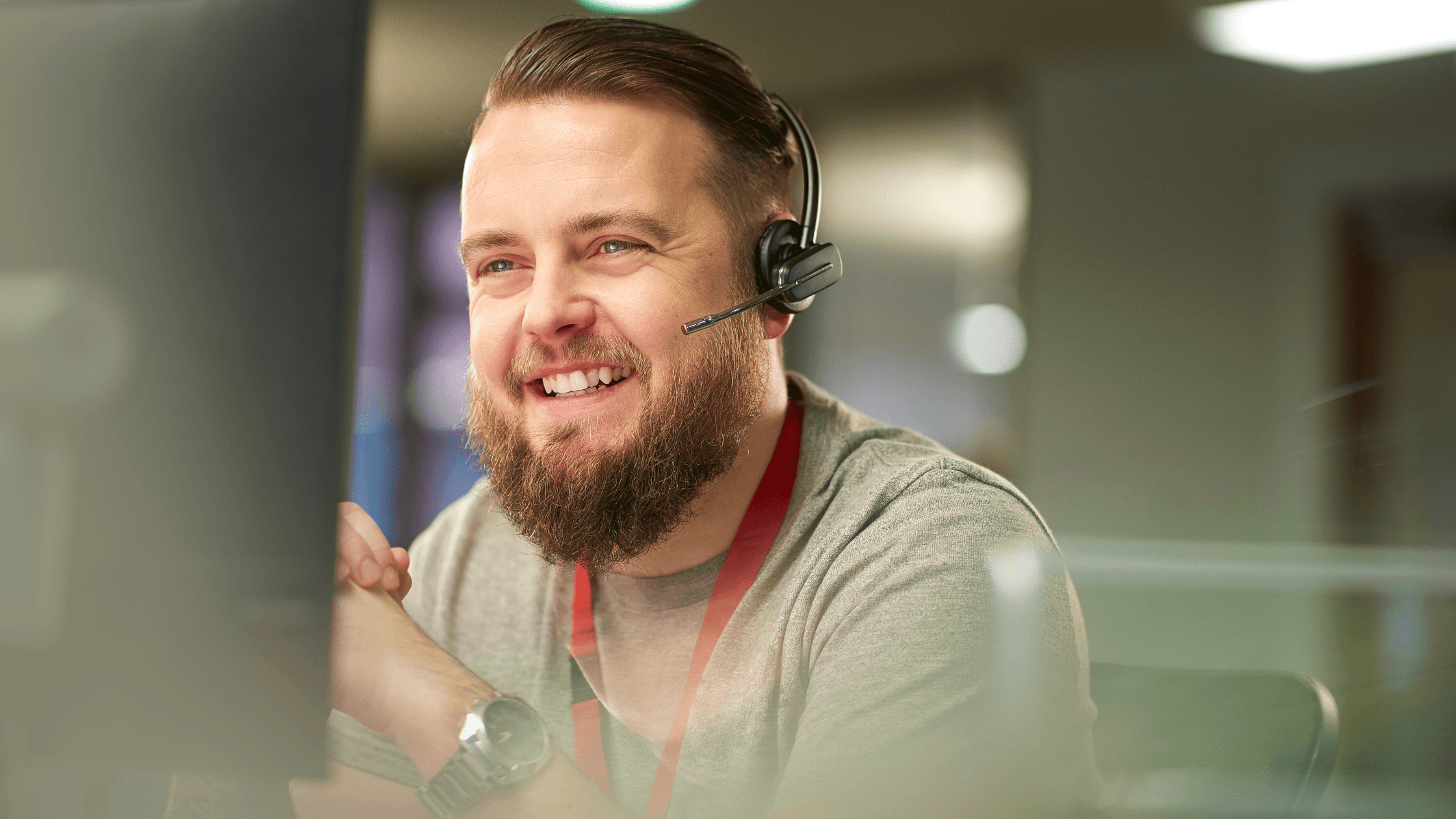 A bearded man wearing a headset and a grey shirt, smiling while working at a computer in an office.
