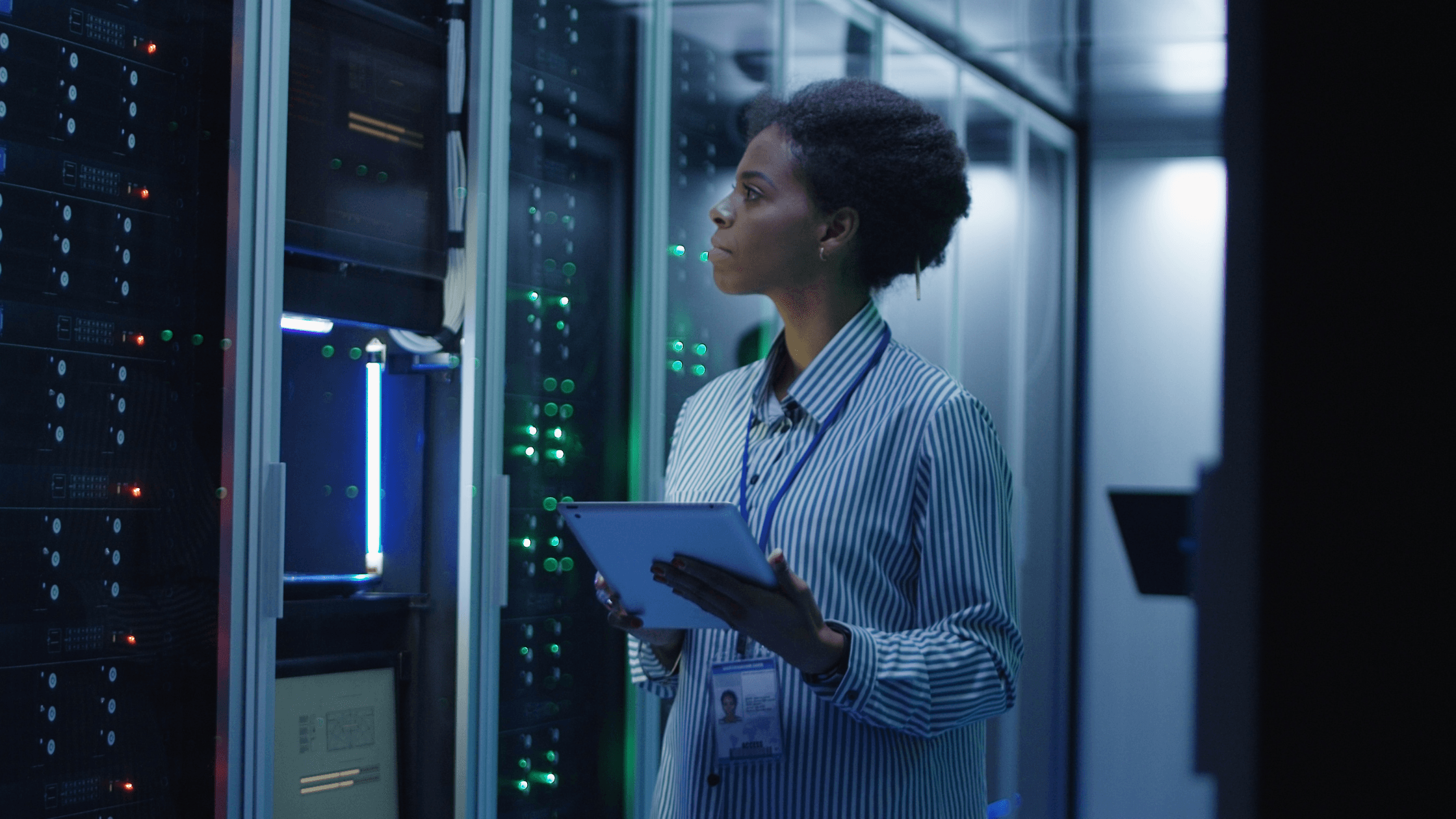A person holding a tablet inspects servers in a dimly lit data center, with glowing lights on the equipment.