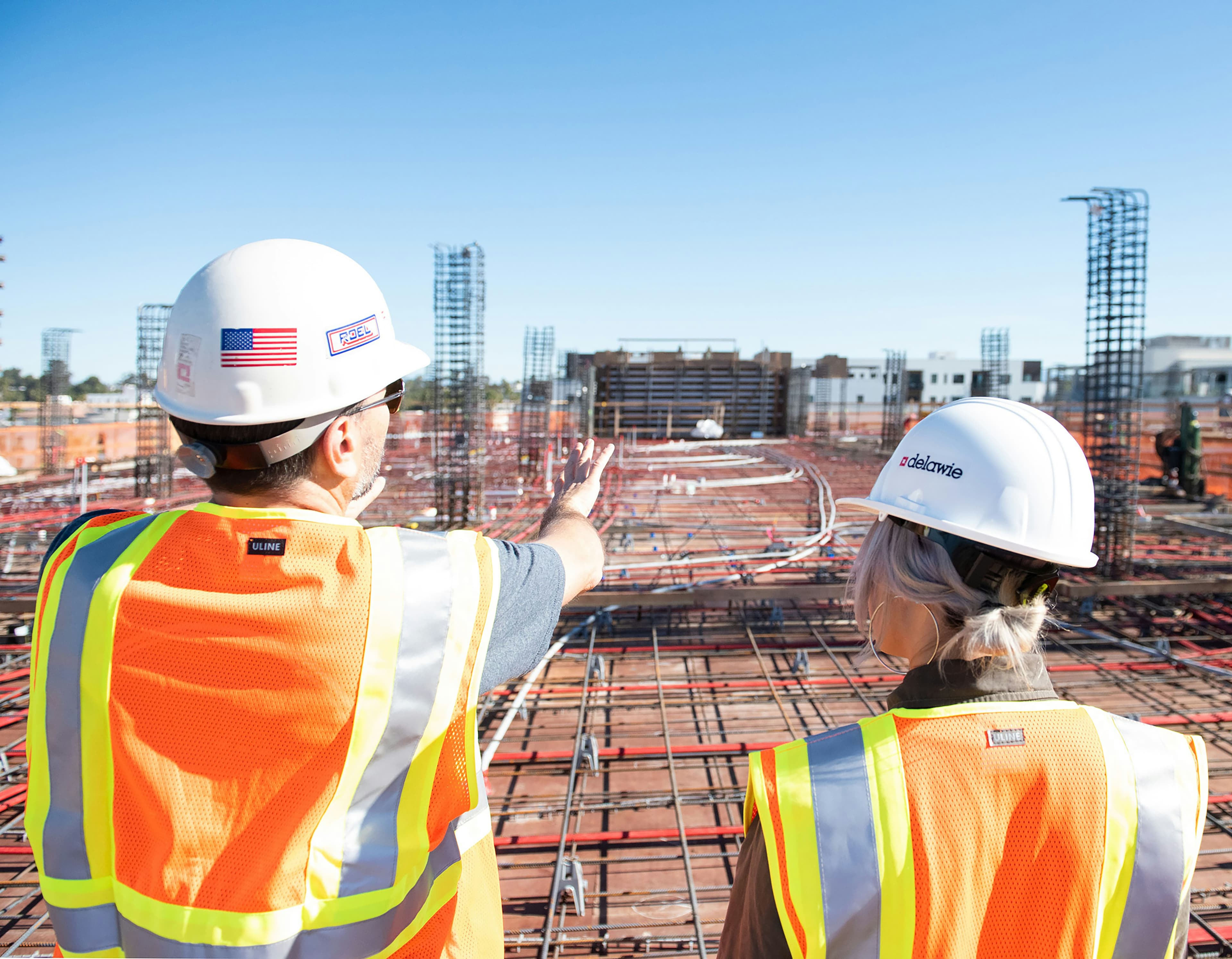 Two construction workers in safety vests and helmets stand on a building site, discussing progress with steel structures visible in the background.