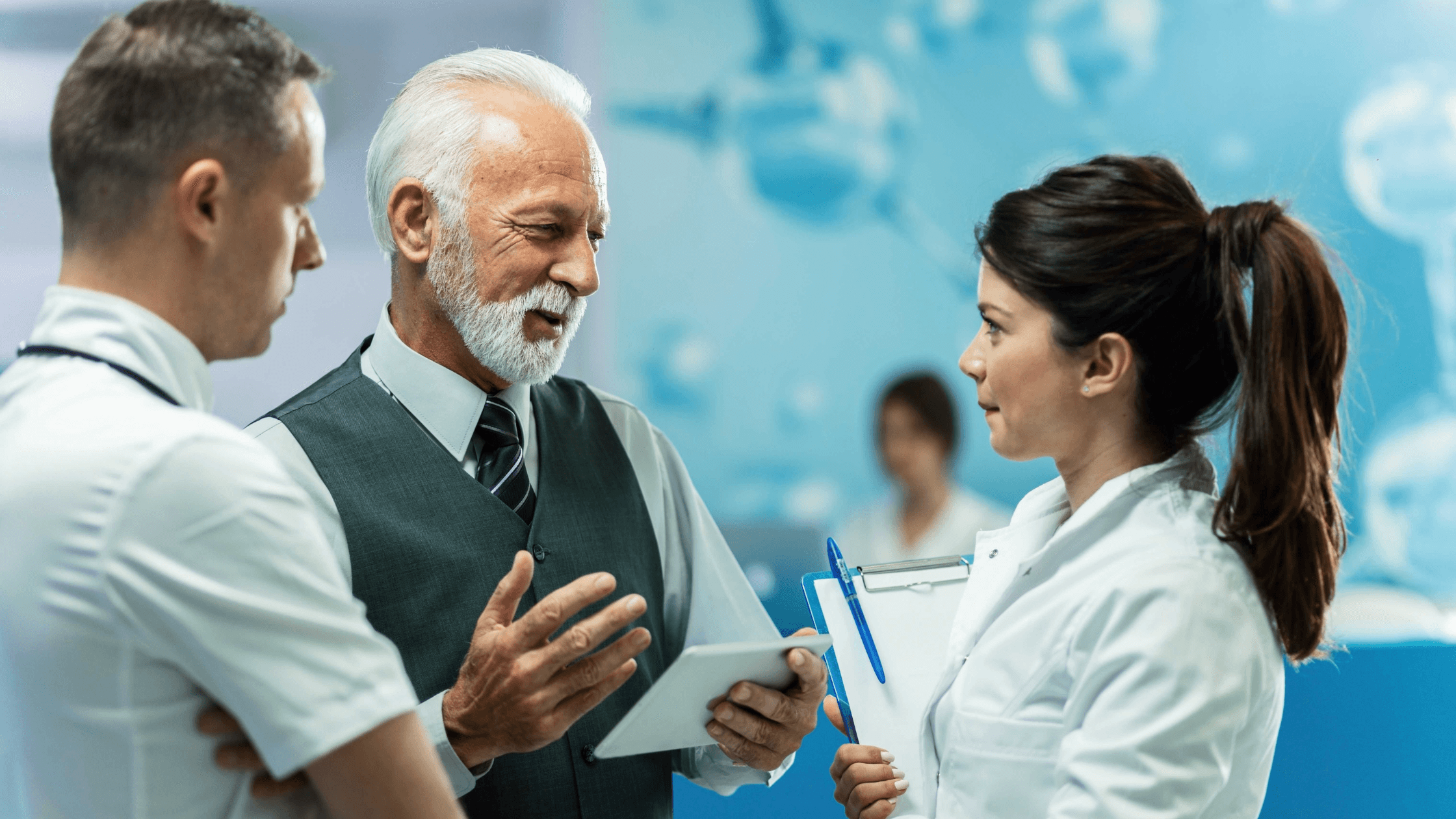 Three medical professionals in discussion; an older man with a tablet and two younger individuals, one holding a clipboard, in a hospital setting.