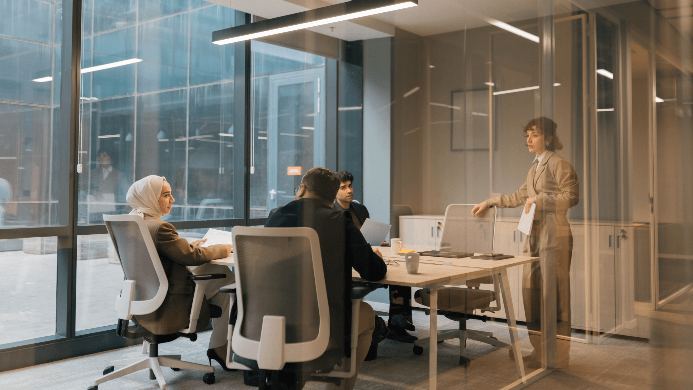 A person stands and presents to three seated colleagues in a modern glass-walled office with large windows and contemporary furniture.