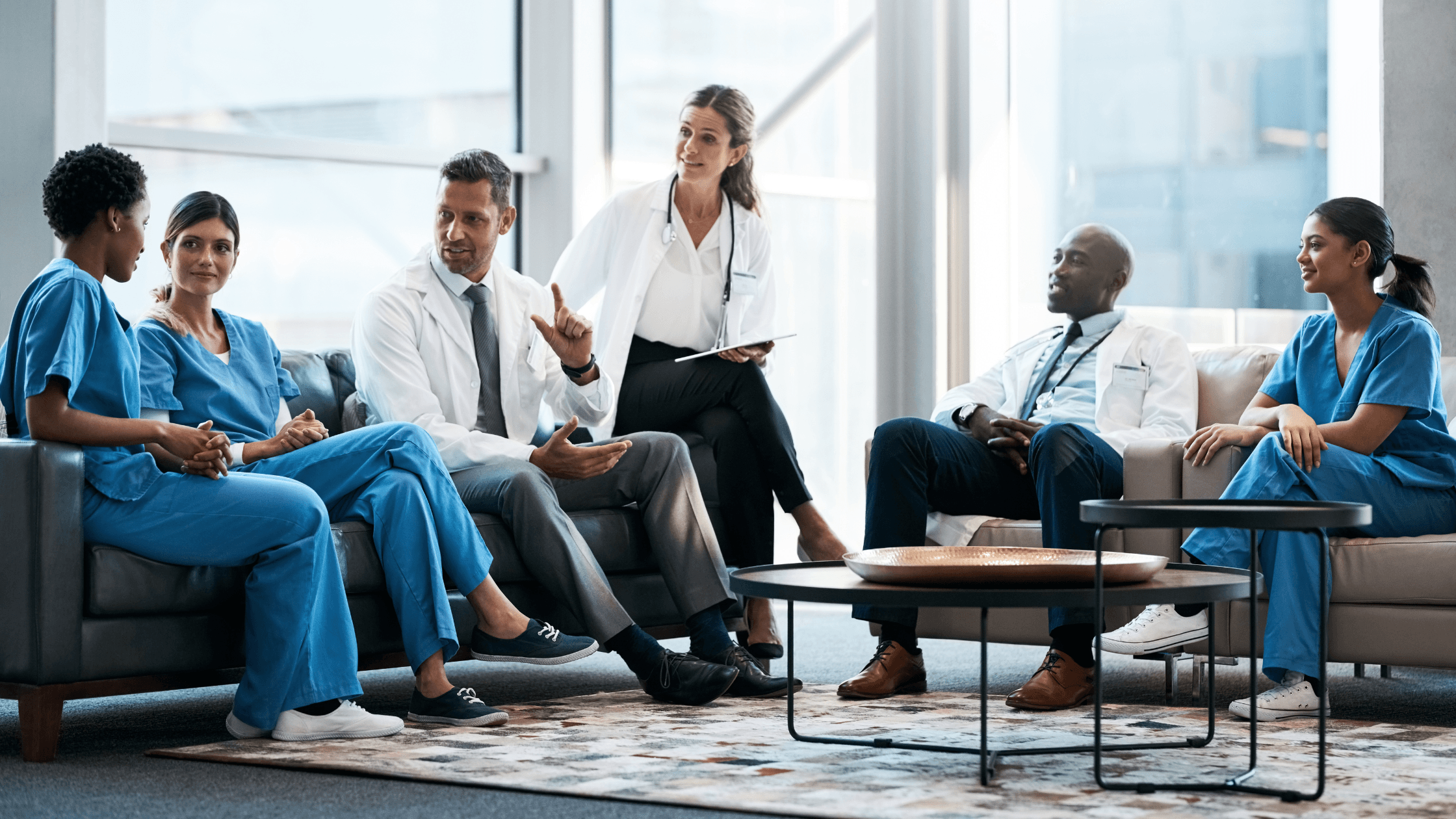 A diverse group of medical professionals in scrubs and lab coats sit and discuss in a modern, well-lit room with large windows.