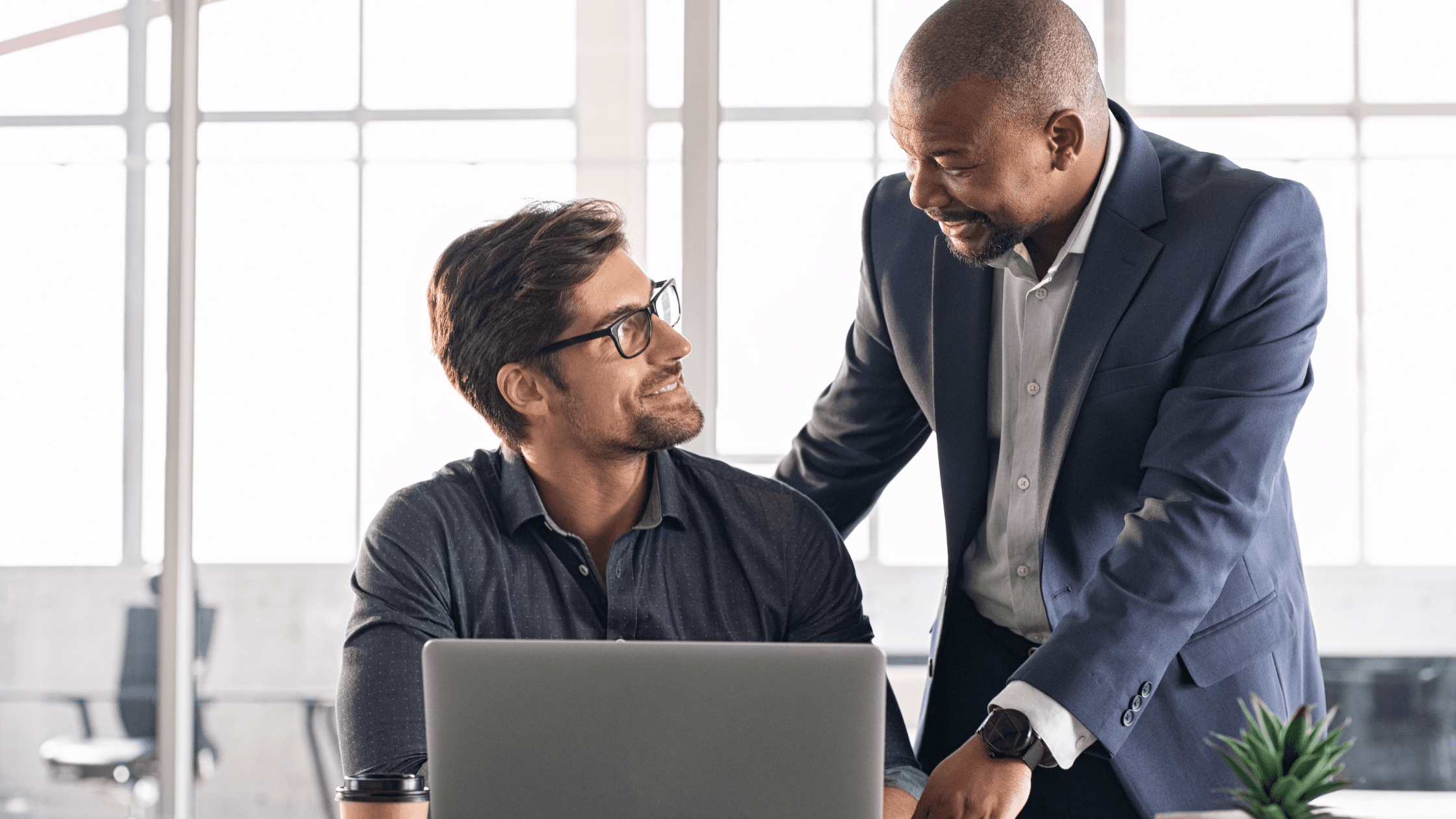 Two men in an office setting, one sitting at a desk with a laptop, and the other standing, smiling and engaged in conversation.