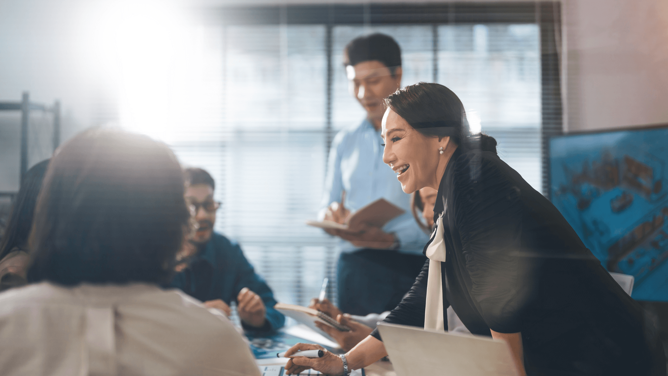A woman leads a meeting, smiling and engaging with colleagues around a table in a bright office.