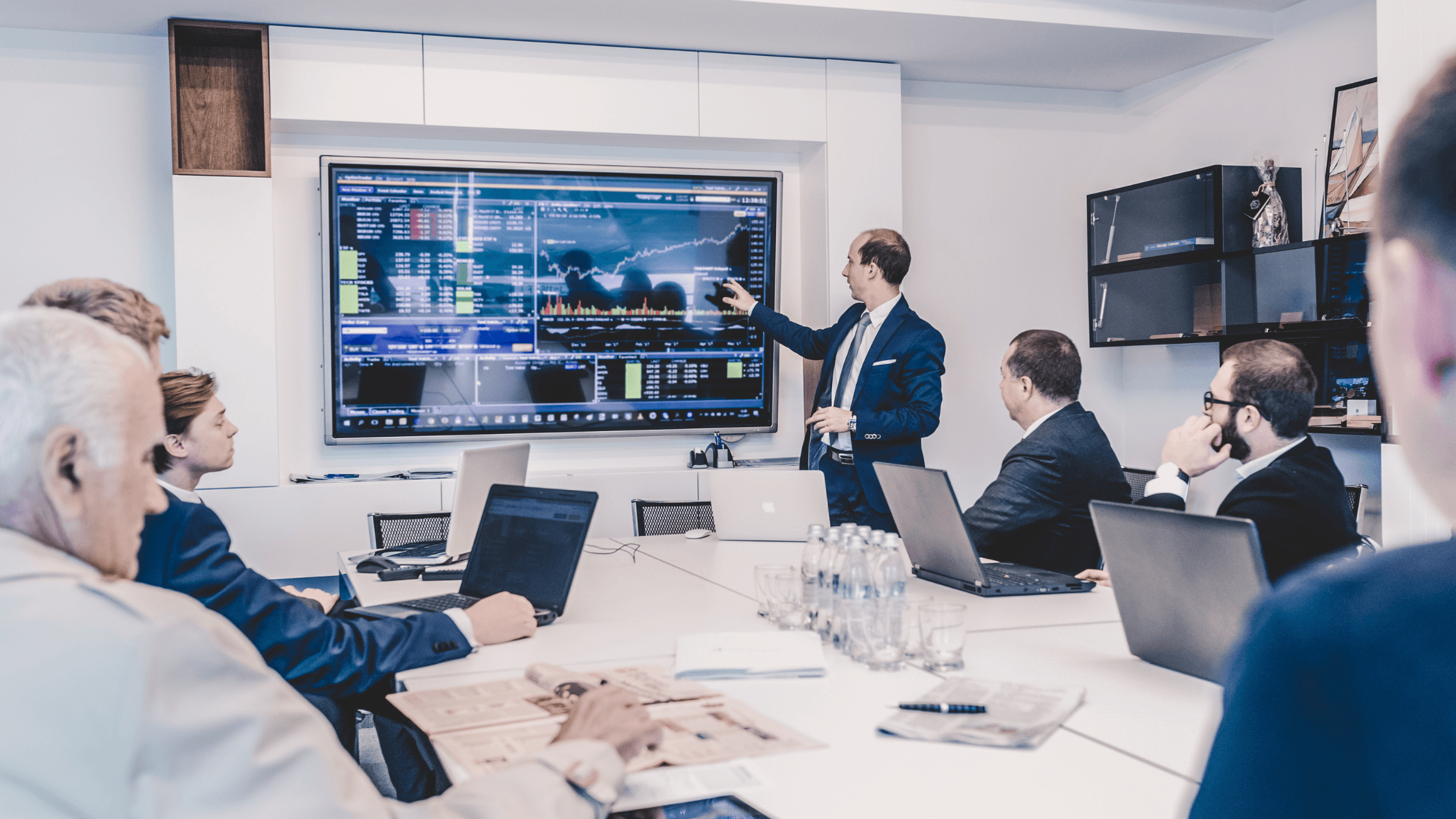 A man in a suit presents financial data on a large screen to a group of colleagues seated around a conference table with laptops.