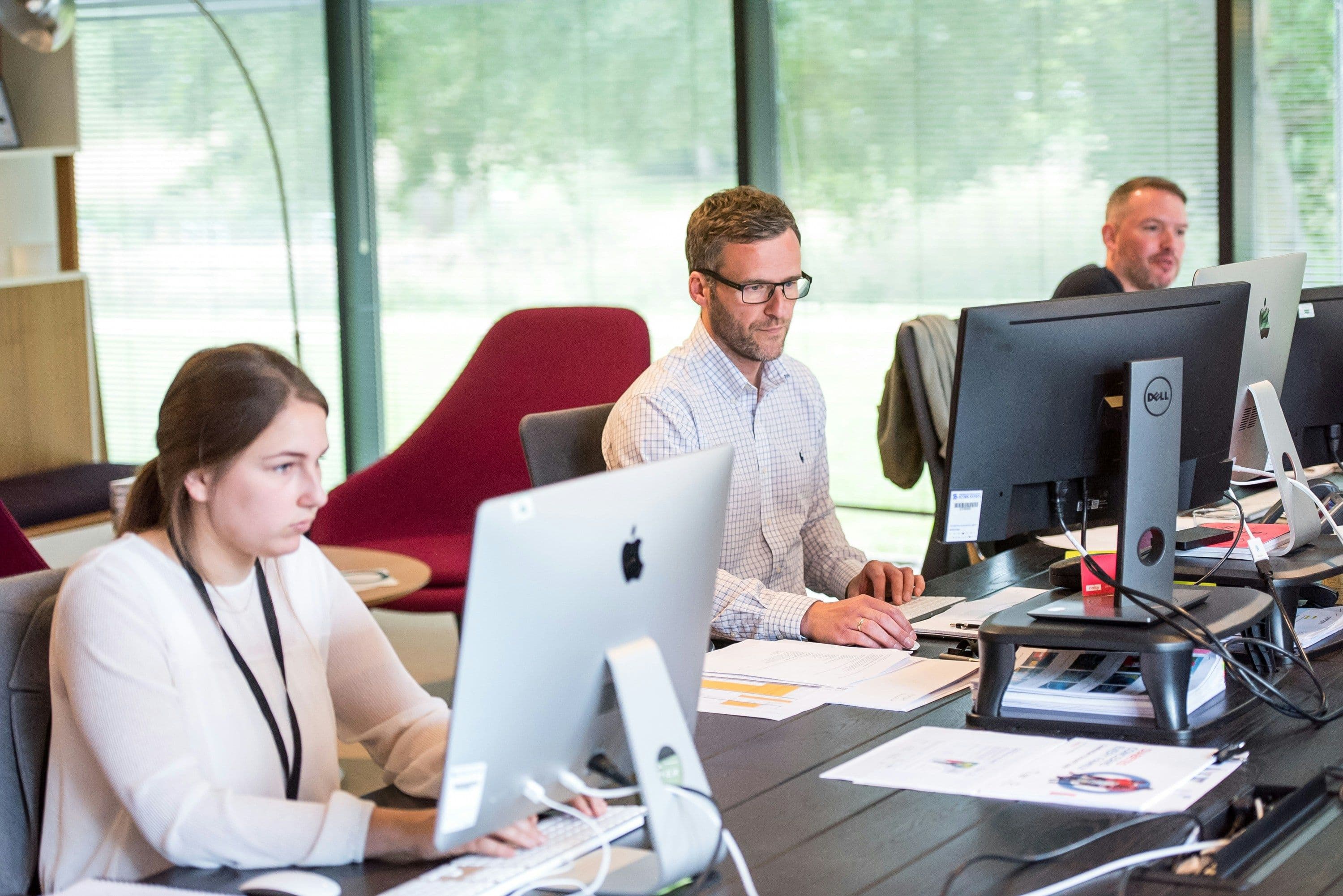 Three people working at computers in a modern office with large windows and natural light.